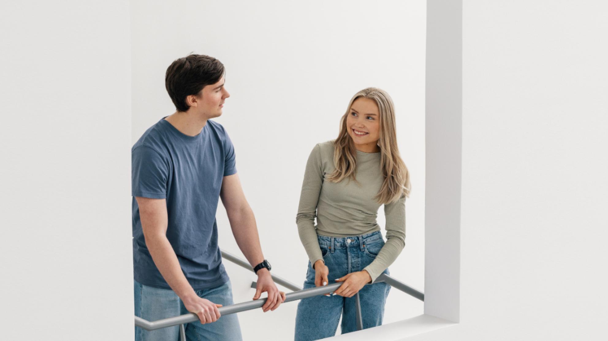 Poika ja tyttö nojaavat porrastasanteella kaiteeseen ja juttelevat keskenään