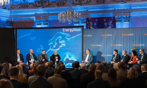 Täysi sali ihmisiä kuuntelee EnergyWeek-tapahtuman paneelikeskustelua lavalla, jossa on useita puhujia istumassa tuoleilla sinistä taustaa vasten. Taustalla näkyy EnergyWeek-logo ja koristeellinen sisätila kattokruunuineen.