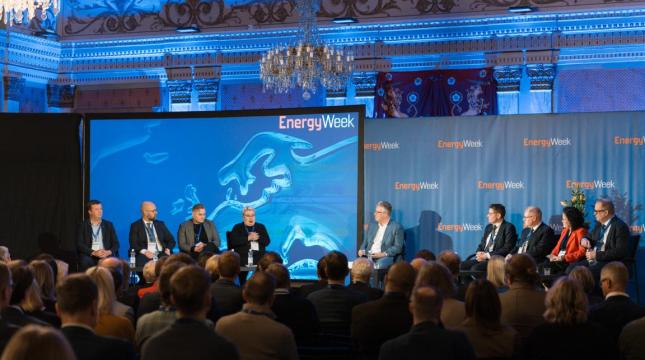 Täysi sali ihmisiä kuuntelee EnergyWeek-tapahtuman paneelikeskustelua lavalla, jossa on useita puhujia istumassa tuoleilla sinistä taustaa vasten. Taustalla näkyy EnergyWeek-logo ja koristeellinen sisätila kattokruunuineen.