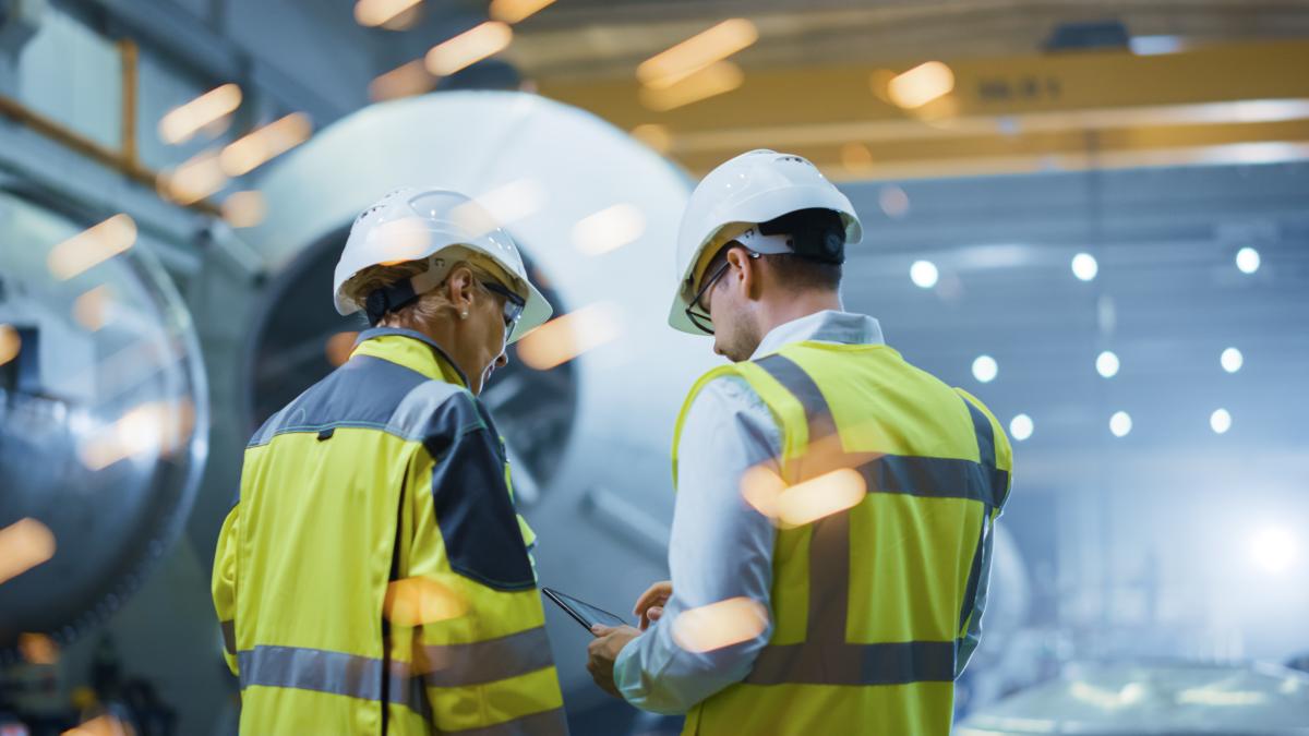 Two heavy industry engineers stand in a pipe manufacturing factory using a digital tablet computer and having a discussion. 